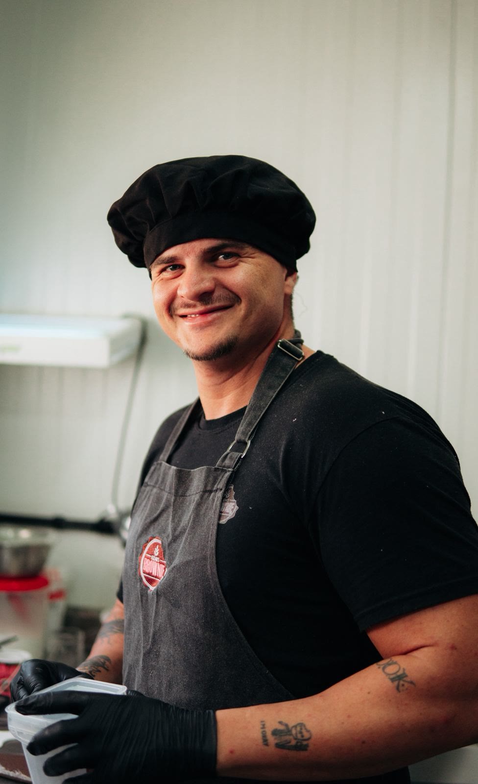 Chef Ale Iuzak sorrindo de touca e avental na cozinha da Iuzaks Brownie